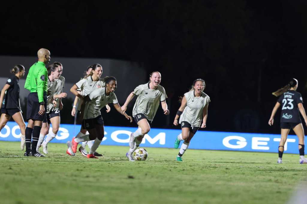 Brooklyn FC women’s players celebrate together on the field at night, running forward with smiles after scoring a goal in the match