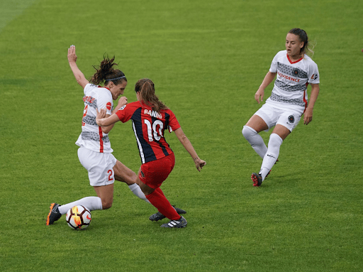 Three women compete for a ball on a soccer pitch